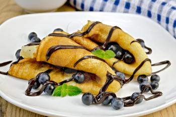 Two pancakes with blueberries, mint and chocolate icing on a white plate, napkin, bowl on the background of wooden boards