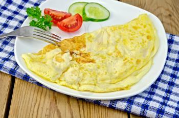 Omelette with slices of tomato, cucumber, parsley and fork on blue checkered napkin against a wooden board