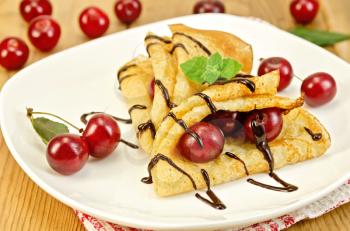Two pancakes with cherries, mint, chocolate icing on a white plate and a napkin on a background of wooden boards