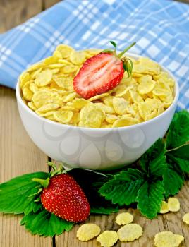 Cornflakes in a white bowl with half a strawberry, leaves and berries of a strawberry, a napkin on a wooden boards background