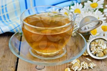 Herbal tea in a glass cup, a metal strainer with dried chamomile flowers, a bouquet of fresh chamomile flowers, napkin against a wooden board