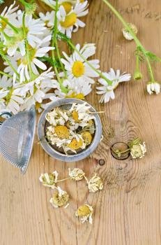Metal strainer with dried chamomile, a bouquet of fresh chamomile flowers on a background of wooden boards