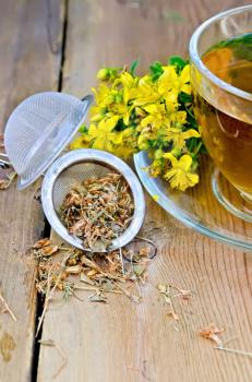 Metal tea strainer with dry flowers tutsan, fresh flowers of Hypericum , tea in a glass cup on a wooden boards background