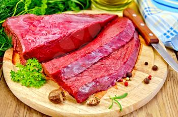 A piece of beef, cut into slices, pepper, parsley, nutmeg and oil in a bottle, napkin, knife on a wooden board