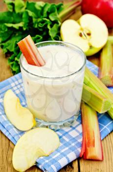 Dairy cocktail in a glass with leaves and stalks of rhubarb, apple slices, blue napkin on a background of wooden boards