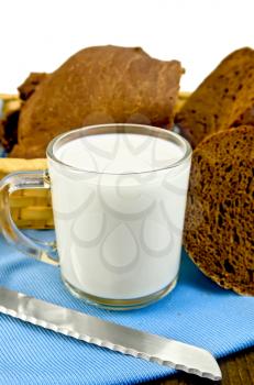Milk in a glass goblet with rye bread in a wicker basket and a knife on a blue napkin and a wooden board isolated on white background