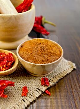 Red pepper powder in a wooden bowl, flake and pods of red pepper in a mortar and a wooden spoon on a background of burlap and wooden board