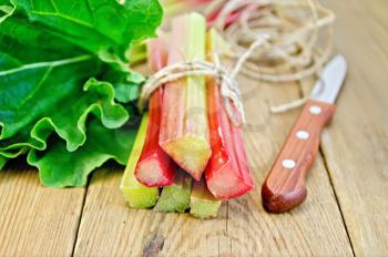 Bundle of stalks of rhubarb with leaf, a knife and a coil of rope on a wooden board