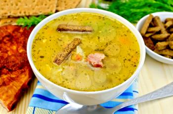 Pea soup with rye croutons in a bowl and on a plate, spoon, pepper, bread, cloth, bacon, dill against a wooden board