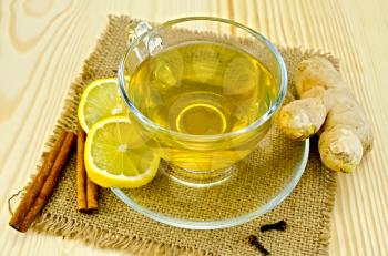 Ginger tea in a glass cup, two slices of lemon, cinnamon, cloves, ginger root on a napkin of burlap and wooden board