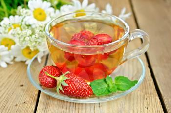 Tea with strawberries in a glass cup, mint, a bouquet of camomiles on a background of wooden boards