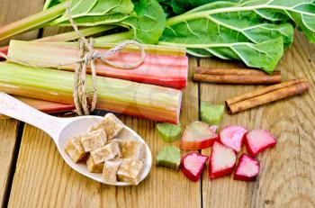 Bundle of stalks rhubarb, cut pieces of rhubarb with a sheet and a knife, a spoon of sugar cubes, cinnamon on a wooden board