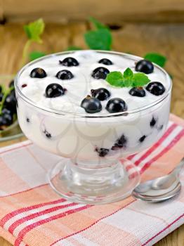 Thick yogurt in glass with black currants and spoon on a napkin, currants in a saucer on the background of wooden boards