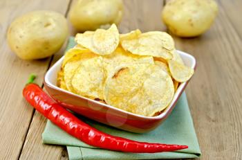 Potato chips in a clay bowl with fresh red chili peppers, fresh potatoes on the background of wooden boards