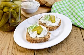 Two sandwiches with cream, pickles and dill, jar of pickles, a napkin on wooden board