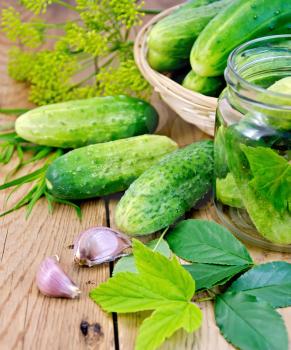 Cucumbers in a glass jar and on the table, garlic, tarragon, dill, cherry leaves and currants on a wooden boards background