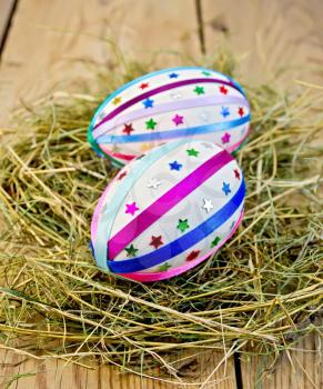 Two easter eggs, decorated with multicolored braid and sparkles as asterisks in the hay on the background of wooden boards
