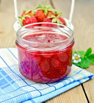 Strawberry jam in a glass jar, strawberries in a wicker basket, strawberry leaves and flowers, napkin on wooden board