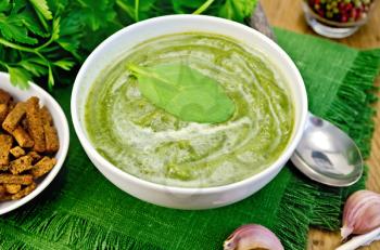 Green soup puree in white bowl with spoon, napkin, parsley, croutons, pepper, garlic on a wooden boards background