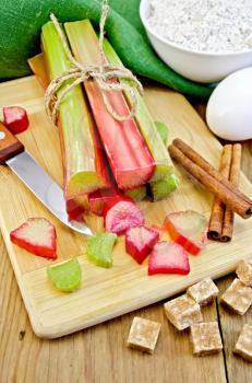 Bundle of stalks rhubarb, cut pieces of rhubarb, knife, sugar cubes, cinnamon, eggs, flour, cloth on a wooden board
