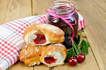 Puff bun with jam, a jar of cherry jam, cherries with leaves, a napkin on a wooden boards background