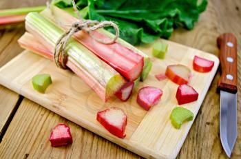 Bundle of stalks rhubarb, cut pieces of rhubarb with a sheet and a knife on a wooden boards background