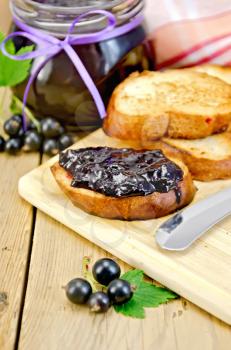 Slices of toasted bread, a glass jar with black currant jam, knife on background wooden board