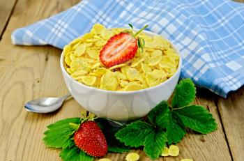 Cornflakes in a white bowl with half a strawberry, leaves and strawberries, napkin on wooden board