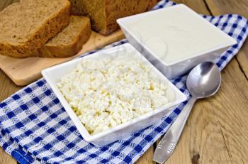 Cottage cheese and sour cream in white square bowls on a checkered blue napkin with spoon, rye bread on a wooden boards background
