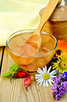 Honey in a glass bowl and a wooden pot, spoon, rosehip berries, flowers, napkin on the background of wooden boards