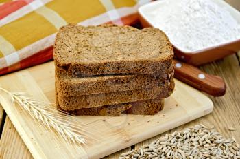 A stack of slices of rye homemade bread with a knife and rye spikelet on plate, napkin, knife, flour and grains on a wooden board