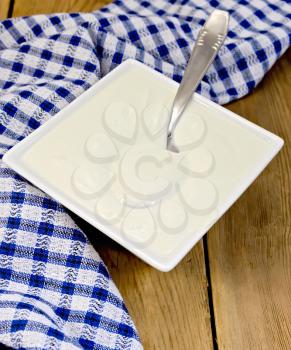 Sour cream in a white square bowl, blue checkered napkin, spoon on a wooden boards background