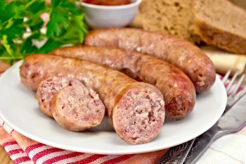 Pork sausages fried in a dish, bread, sauce, tomato, parsley, fork, knife, napkin on the background of wooden boards