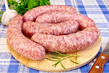 Raw pork sausages on a round wooden board, knife, rosemary, parsley and garlic on blue background checkered cloth