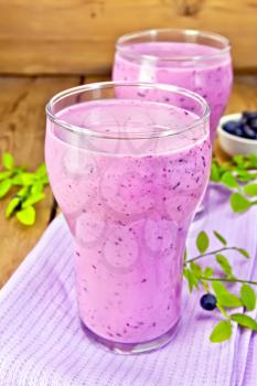 Two tall glass of milkshake of blueberries on a lilac napkin, blueberries on a wooden boards background