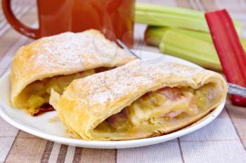 Strudel with rhubarb and fork on a plate, mug, rhubarb stalks on linen tablecloth background