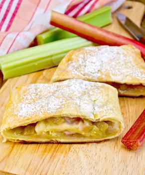 Strudel with rhubarb, knife, napkin, rhubarb stalks on the background of wooden boards