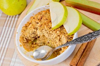 Crumble with pears and rhubarb in a white bowl on a wooden board, rhubarb stalks and pear, cinnamon, spoon on a background of linen tablecloth