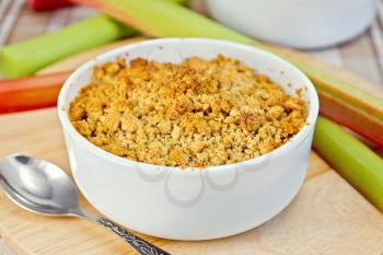 Crumble with rhubarb in a white bowl on a wooden board, rhubarb stalks, spoon on a background of a linen tablecloth