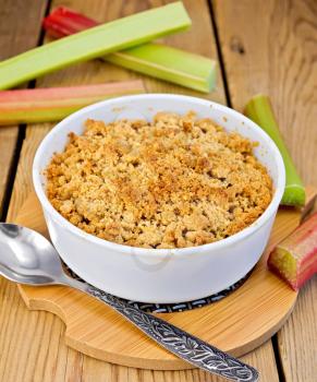 Crumble with rhubarb in a white bowl, rhubarb stalks, spoon on a background of wooden boards