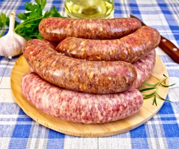 Raw pork and beef sausages on a round wooden board, knife, rosemary, parsley and garlic on a background of blue plaid fabric
