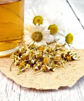 Dried chamomile flowers on paper, tea in glass mug, fresh daisy flowers on a background of pale wooden plank