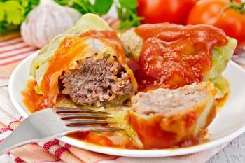Stuffed cabbage meat in cabbage leaves with tomato sauce and a fork on a plate on a napkin, tomatoes, parsley and garlic on a background of pale wooden plank