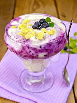 Dessert milk with blueberry, cornflakes, curd, spoon in a glassware on a purple napkin on a wooden boards background