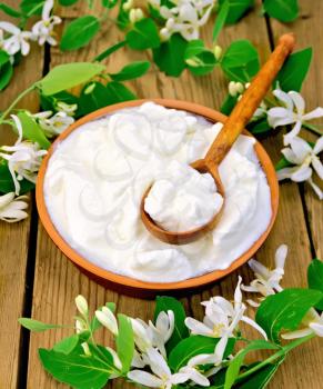 Thick yogurt in a clay bowl with spoon and white flowers of honeysuckle on a wooden boards background