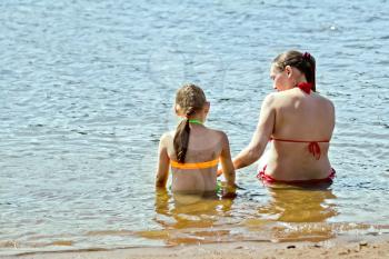 Mother and daughter sitting in the water near the sandy shore