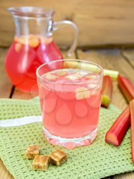 Compote from rhubarb in glassful and pitcher, sugar, rhubarb stalks, napkin on a wooden boards background