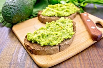 Two slices of rye bread with guakomole avocado on a plate, napkin, parsley on a wooden boards background