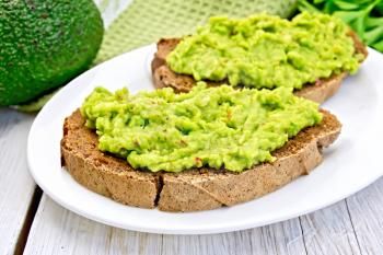 Two slices of rye bread with guakomole avocado on a plate, napkin, parsley on the background light wooden boards