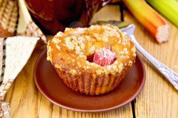 Cupcake with rhubarb on a plate, mug, brown napkin, rhubarb stalks on a wooden boards background
